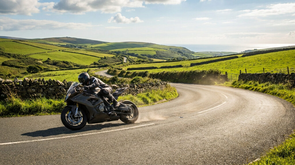 Moto sportive noire penchée dans un virage sur une route sinueuse verdoyante. Un pilote en tenue de course soulève de la poussière. Mer à l'horizon.